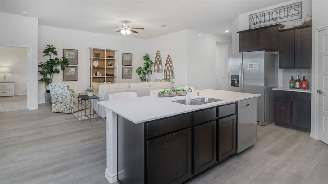 Open-concept living space featuring wood-finish flooring, a kitchen island with an integrated sink, stainless steel appliances, dark wood cabinetry, and a ceiling fan