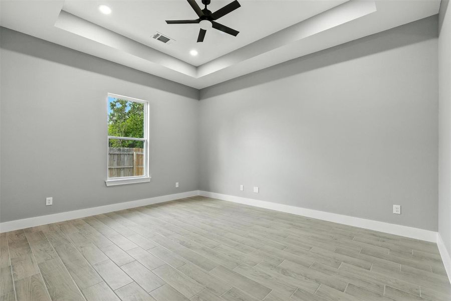 This photo showcases a spacious, modern room with light gray walls and wood-look tile flooring. The room features a tray ceiling with recessed lighting and a sleek ceiling fan. A window provides natural light and views of the fenced backyard.
