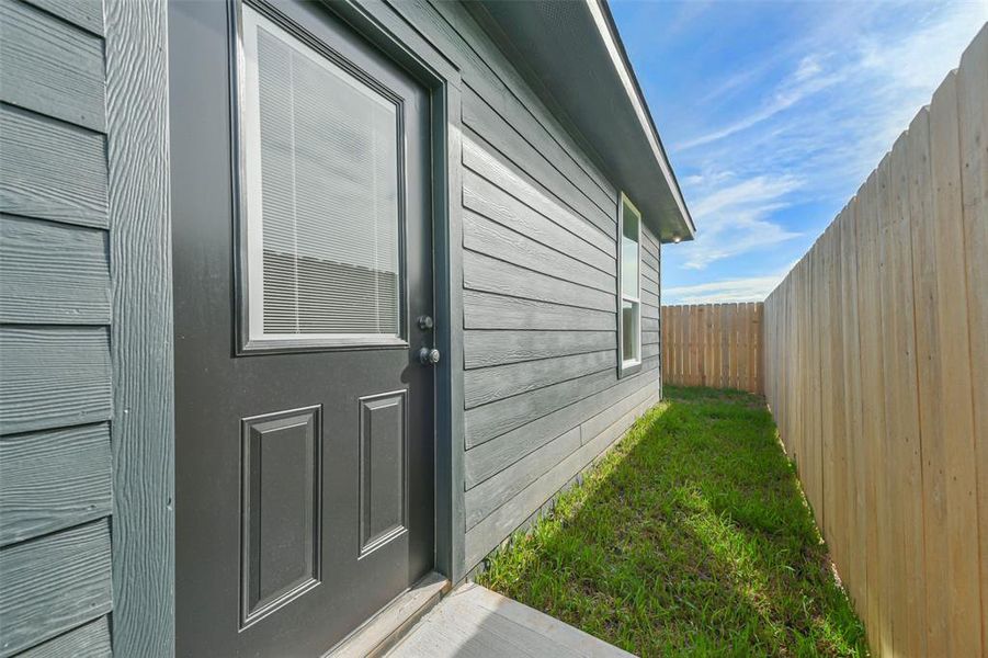 This image shows the side exterior of a modern home with gray siding, featuring a black door and a single window. The property includes a neatly maintained, narrow strip of lawn and is enclosed by a tall, wooden privacy fence.