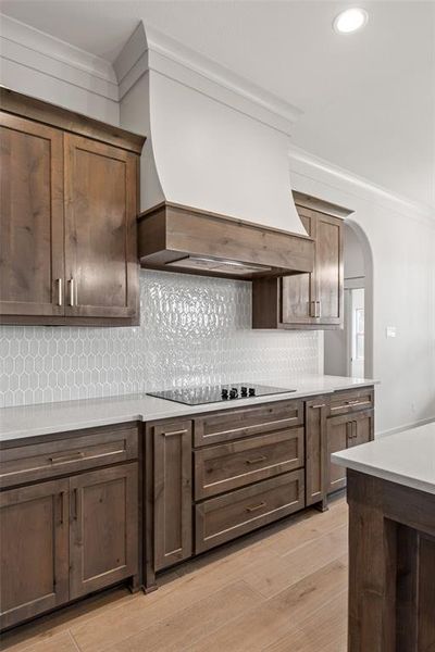 Kitchen featuring custom wood cabinetry, a built-in cooktop, light-toned countertops, decorative tile backsplash, and a custom range hood with crown molding accents