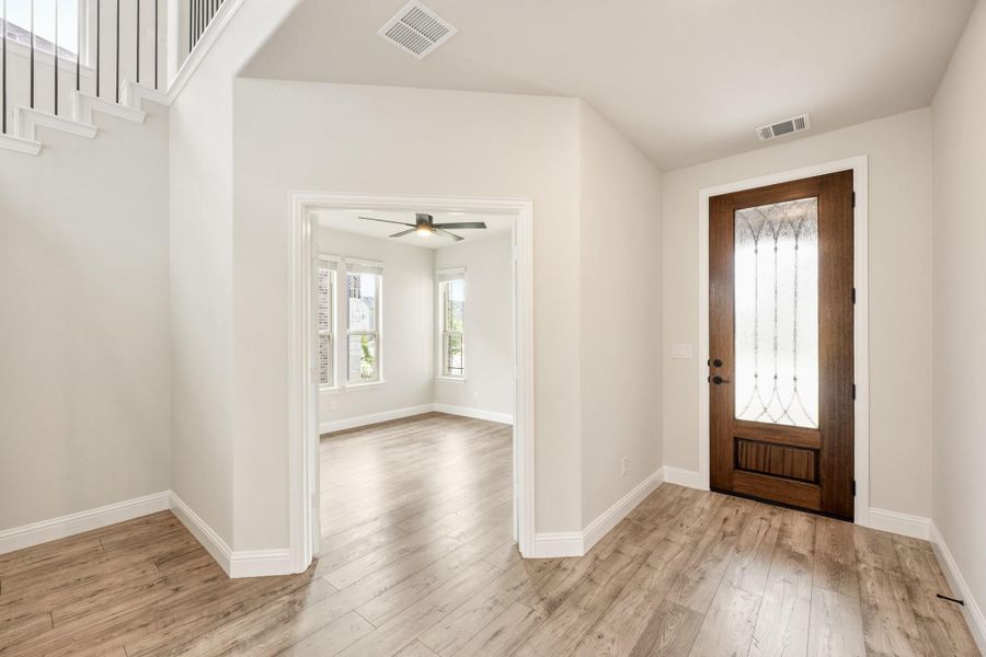 Entryway with wood front door, decorative glass panel, and light hardwood floors leading to an open room with ceiling fan.