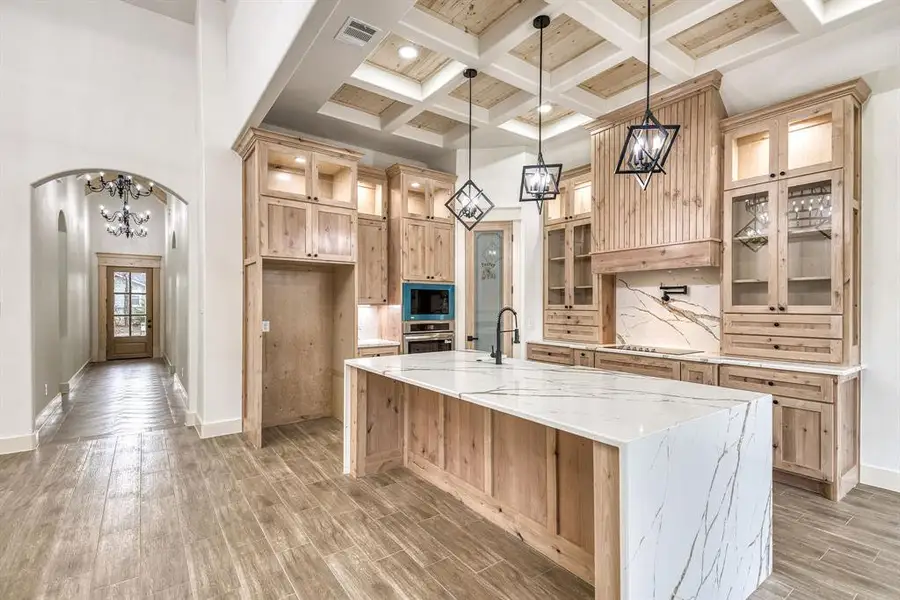 Kitchen featuring light wood finish cabinets, light stone countertops, hanging lights, glass insert cabinets, and coffered ceiling