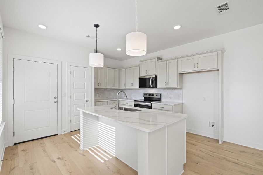 Kitchen featuring appliances with stainless steel finishes, pendant lighting, light stone countertops, a kitchen island with sink, and light wood finished floors