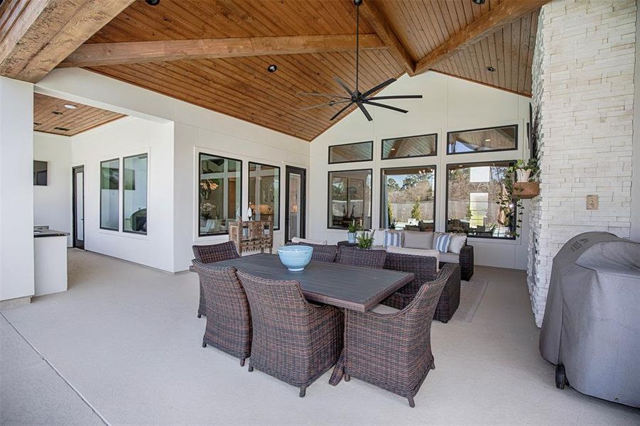 This photo showcases a spacious, covered patio with a vaulted wooden ceiling and modern design. It features a dining area with wicker chairs, a cozy seating area, and a built-in grill. Large windows provide ample natural light, enhancing the inviting outdoor living space.