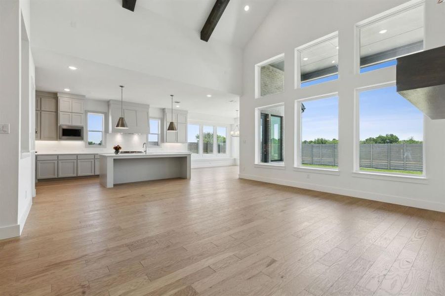 Open-concept living area featuring a vaulted ceiling with exposed dark wood beams, light wood-finish flooring, a wall of six large windows, and a modern kitchen Open-concept living area featuring a vaulted ceiling with exposed dark wood beams, light wood-finish flooring, a wall of six large windows, and a modern kitchen
