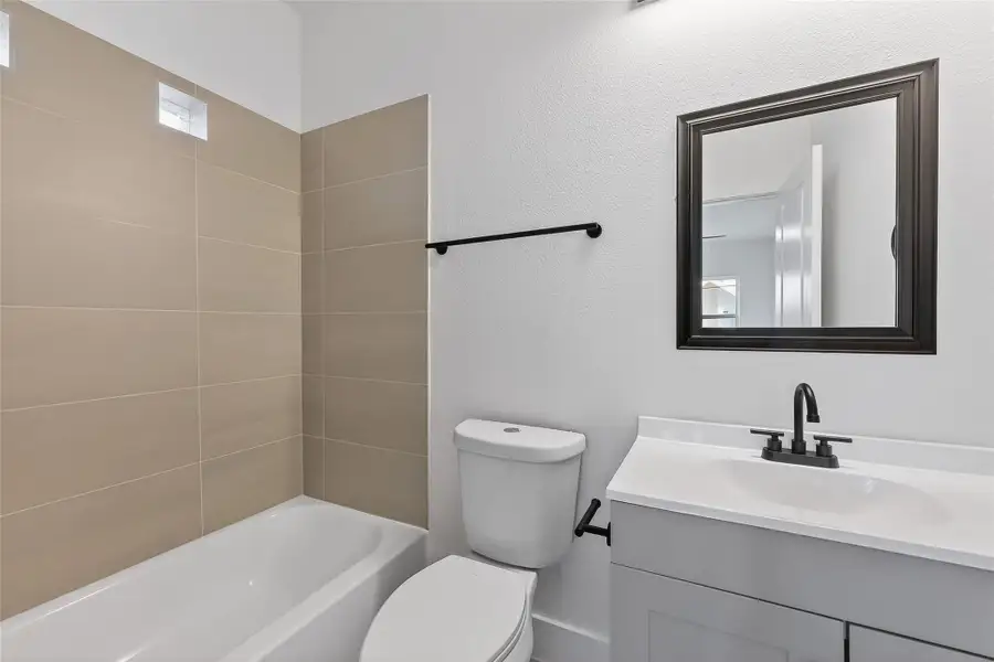 This bathroom features a modern design with neutral beige tiles in the shower area, a sleek white vanity, and a black framed mirror. The fixtures are contemporary, and the space is well-lit, providing a clean and inviting atmosphere.
