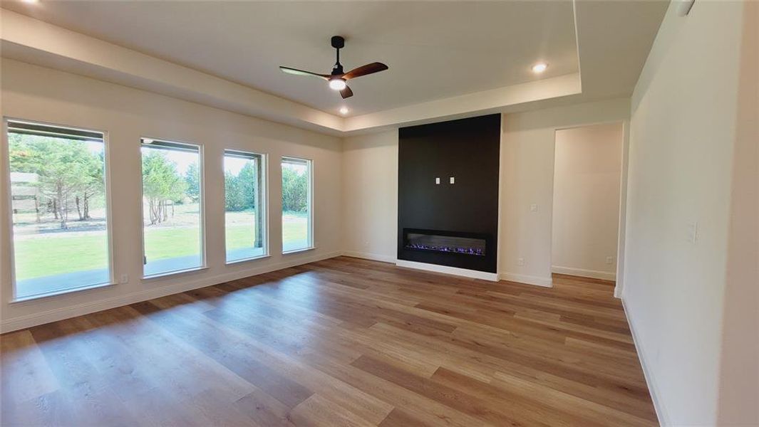 Spacious room featuring wood-finish flooring, a contemporary electric fireplace with a dark accent wall, a tray ceiling with recessed lighting, and a ceiling fan