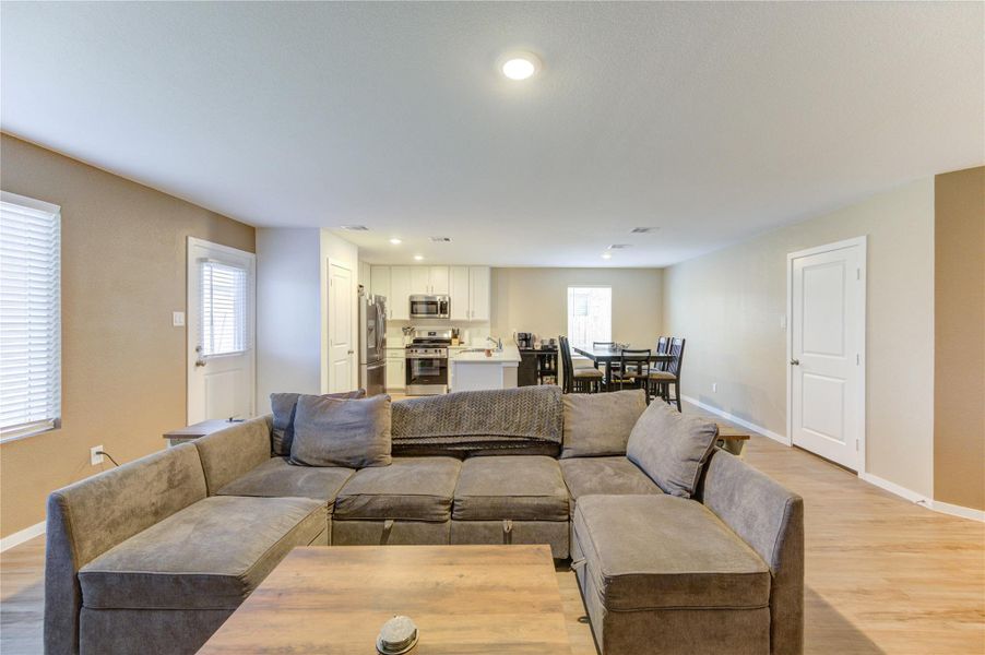 This photo shows an open-concept living space with a cozy sectional sofa and a wooden coffee table. The room flows into a modern kitchen equipped with stainless steel appliances and an adjacent dining area. The space is well-lit with recessed lighting and natural light from windows, creating a warm and inviting atmosphere.