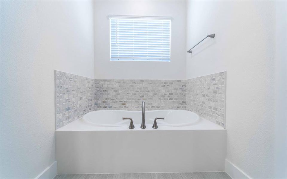 Soaking tub with a mosaic tile surround, featuring a window with blinds, a wall-mounted towel bar, and wood-finish flooring