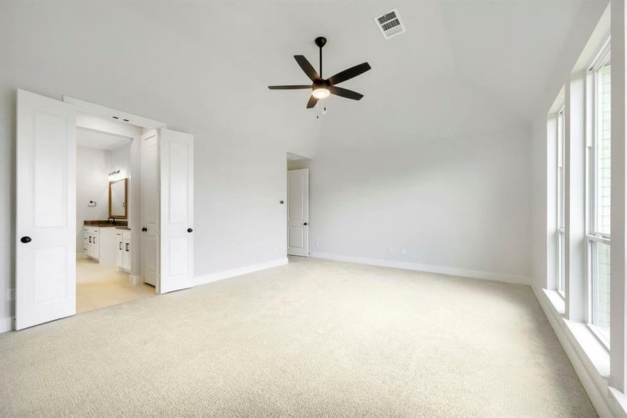 Spacious carpeted room featuring a vaulted ceiling with a ceiling fan, multiple windows, and double doors opening to a bathroom with a vanity