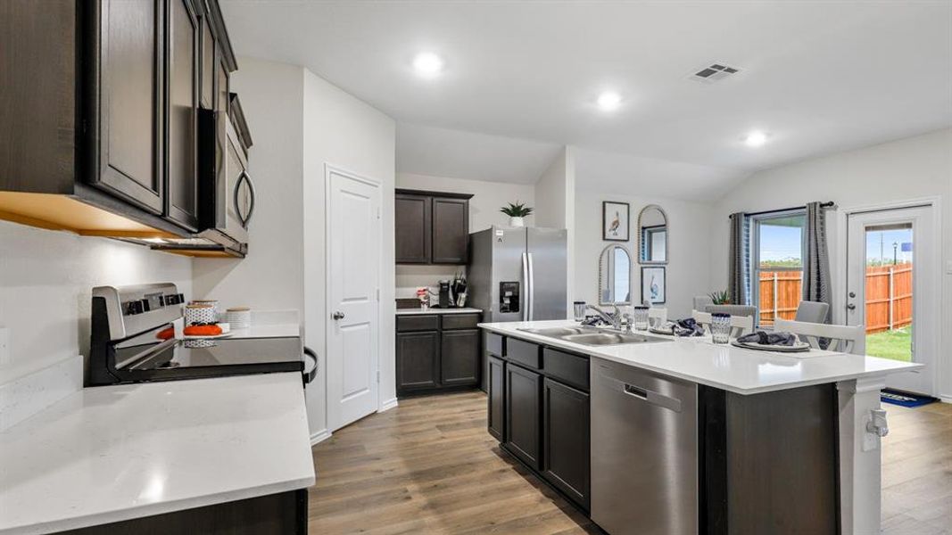 Kitchen featuring stainless steel appliances, vaulted ceiling, light wood finished floors, light countertops, and a kitchen island with sink Kitchen featuring stainless steel appliances, vaulted ceiling, light wood finished floors, light countertops, and a kitchen island with sink