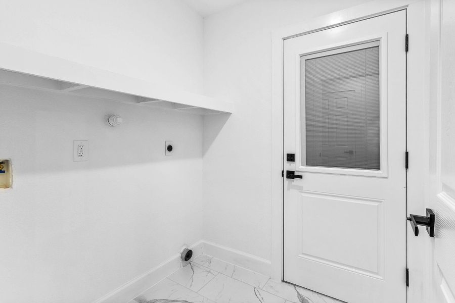 This photo shows a small, bright laundry room with white walls and a door featuring built-in blinds. It includes hookups for a washer and dryer, a shelf for storage, and tiled flooring.