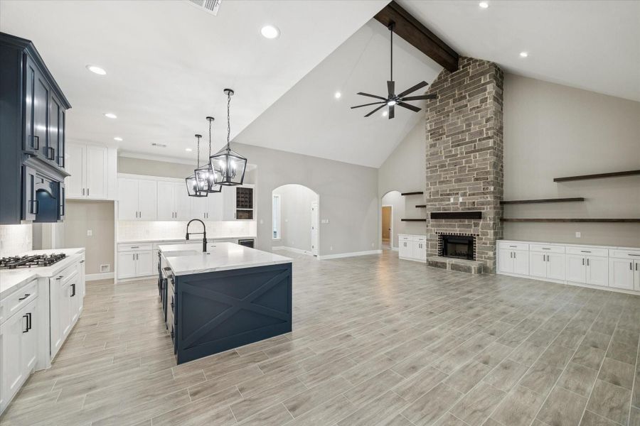 View from dining room toward family room.  Vaulted 20ft. beam ceiling.  Lots of built-in closed cabinets for storage.  Quartz counters with lots of counter space and floating shelves above.  Drop lighted ceiling fan.   Designer blue island and upper cabinets above the cook top.  Arched opening in the middle of the photo leads to the entry hall. Arched opening on the right leads to the secondary bedrooms and baths.