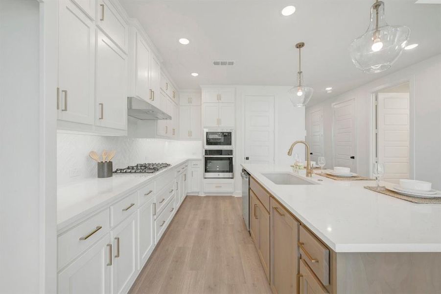 Bright kitchen featuring white shaker cabinetry, an integrated gas range, built-in double ovens, a central island with an undermount sink, and wood-finish flooring