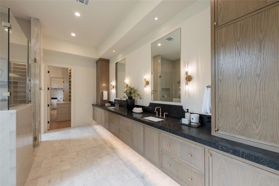 This angle reveals the full expanse of the primary bath’s custom double vanity—a rich mix of craftsmanship and materiality. The textured oak cabinetry anchors the space with a warm, organic feel, while the honed black countertops add depth and contrast. Oversized mirrors, flanked by elegant glass sconces in a brass finish, reflect the thoughtful balance of form and function. This angle reveals the full expanse of the primary bath’s custom double vanity—a rich mix of craftsmanship and materiality. The textured oak cabinetry anchors the space with a warm, organic feel, while the honed black countertops add depth and contrast. Oversized mirrors, flanked by elegant glass sconces in a brass finish, reflect the thoughtful balance of form and function.