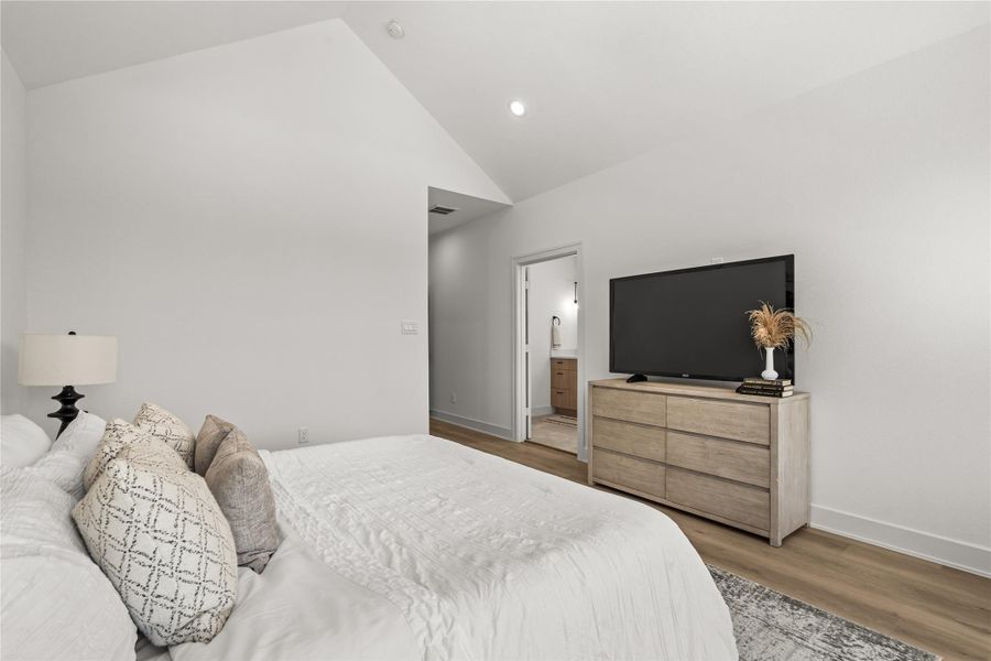 This view from the windows shows the smooth transition from the bed to the rest of the suite. The wide layout provides a clear path toward the attached primary bathroom, while a low-profile media console opposite the bed keeps the room clean and clutter-free.
