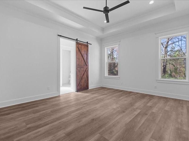 Empty room featuring a tray ceiling, light wood finished floors, a barn door, crown molding, and a ceiling fan