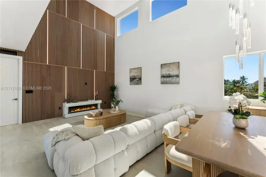 Double-height ceilings with clerestory windows, vertical slat wood-finish accent wall with integrated lighting, built-in linear electric fireplace, stone-finish flooring, and multi-light hanging chandelier Double-height ceilings with clerestory windows, vertical slat wood-finish accent wall with integrated lighting, built-in linear electric fireplace, stone-finish flooring, and multi-light hanging chandelier