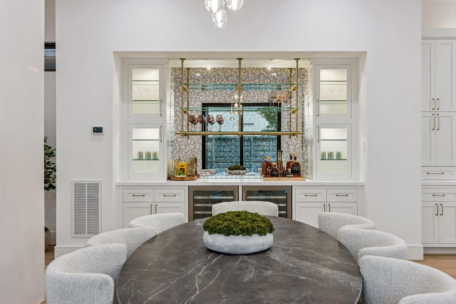 The back wall showcases a luxurious built-in wine display and bar area with illuminated shelving, glass fronted doors. The center of the wine display features Antique mirrored backing with Iron Abode Leo Shelving Unit finished is Brushed Brass. Integrated wine refrigerators below the counter.