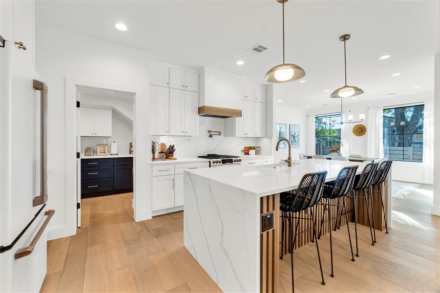 Kitchen with white cabinetry, high end fridge, light wood finished floors, decorative light fixtures, and a large island with sink Kitchen with white cabinetry, high end fridge, light wood finished floors, decorative light fixtures, and a large island with sink