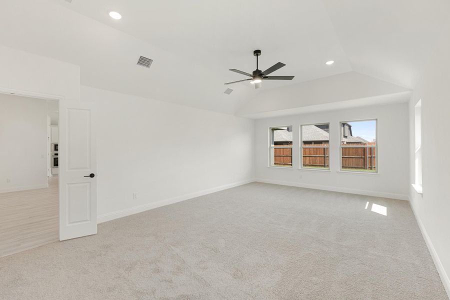 Empty bedroom with vaulted ceiling, ceiling fan, carpet flooring, and three windows overlooking backyard fence