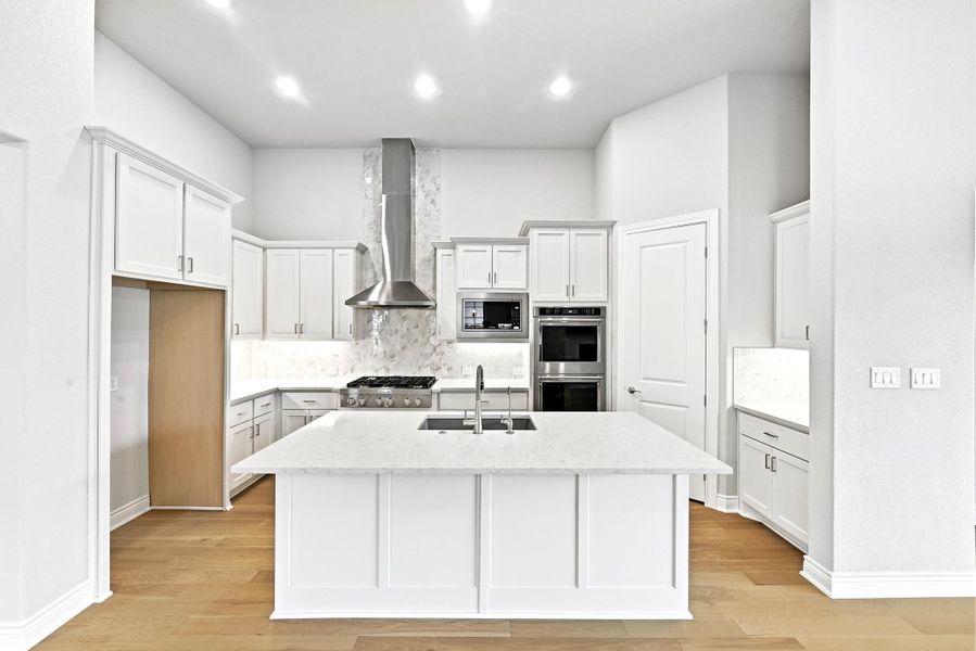 Modern kitchen featuring an expansive center island with an undermount sink, white shaker cabinetry, stainless steel appliances including a gas range and double ovens, and wood-finish flooring