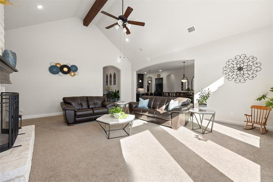 Living room with ceiling fan, carpet flooring, a stone fireplace, high vaulted ceiling, and beamed ceiling Living room with ceiling fan, carpet flooring, a stone fireplace, high vaulted ceiling, and beamed ceiling