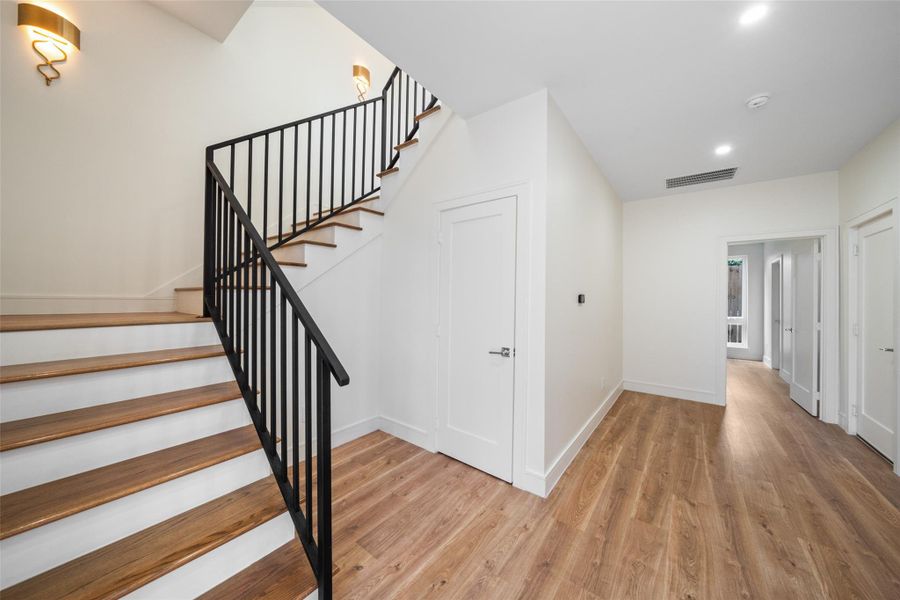 Modern staircase with sleek black railings and wooden steps, leading up from a bright, spacious hallway with light wood flooring and white walls. The space is well-lit with recessed lighting and contemporary wall fixtures, creating a warm and inviting entryway.