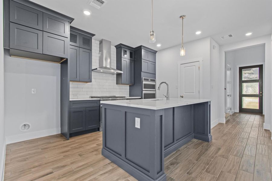 Kitchen with wall chimney exhaust hood, backsplash, hanging light fixtures, a kitchen island with sink, and light stone counters