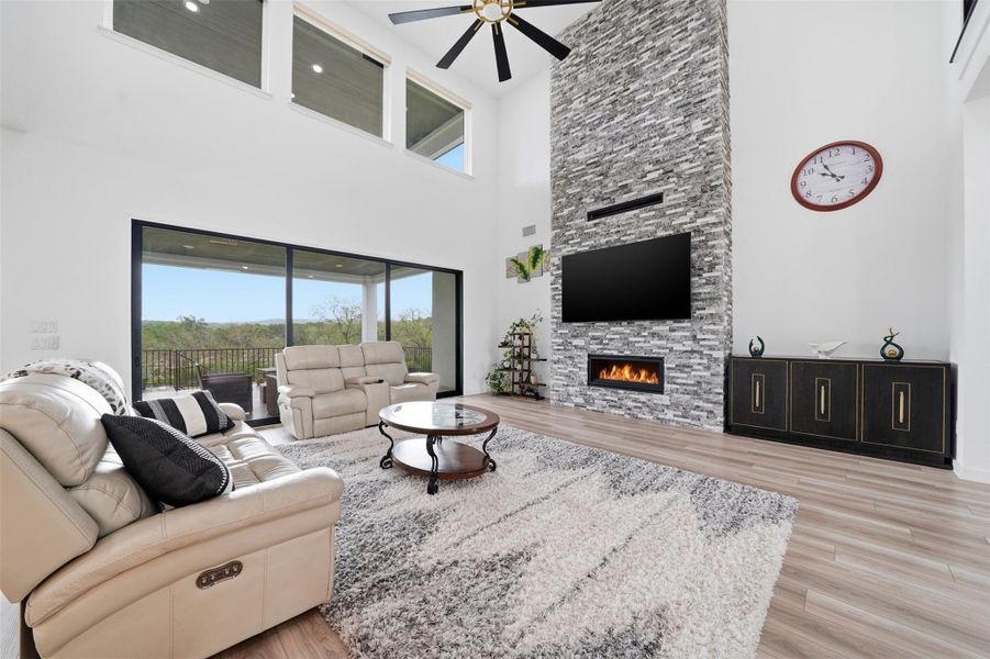 Living room with high ceilings, featuring a floor-to-ceiling stone-clad fireplace, a ceiling fan, and large windows with views of the outdoor landscape