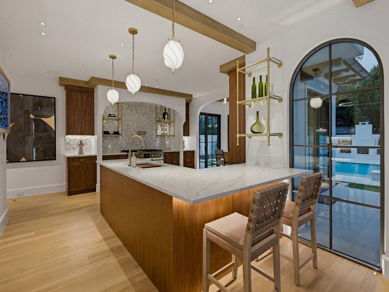 Kitchen featuring a large central island with a light-toned countertop, wood-finish cabinetry, a patterned tile backsplash, and a gold-toned pot filler