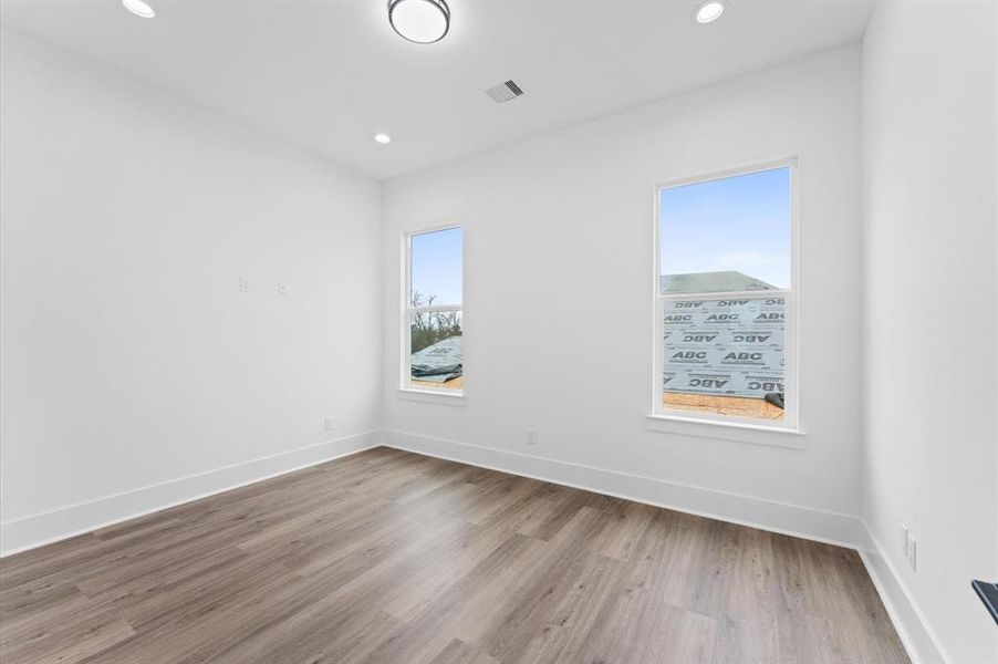 This photo shows a bright, empty room with clean white walls and two large windows. The room features wood-look flooring and recessed lighting, providing a modern and fresh look.