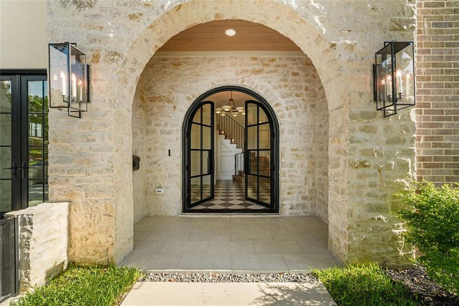 Stone archway entry featuring a wood-paneled ceiling, black metal-framed glass doors, and two contemporary outdoor sconces