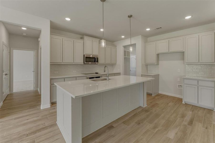 Kitchen featuring a large central island with an integrated sink, light-toned cabinetry, light countertops, subway tile backsplash, and wood-finish flooring
