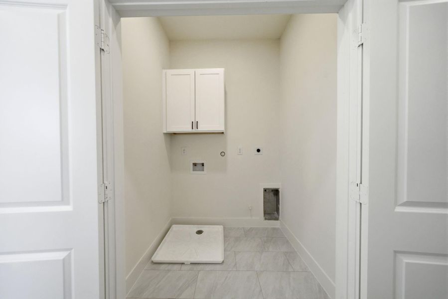 This photo shows a spacious laundry room with tiled flooring, gas/electric hookups for a washer and dryer, and a small cabinet for storage above.