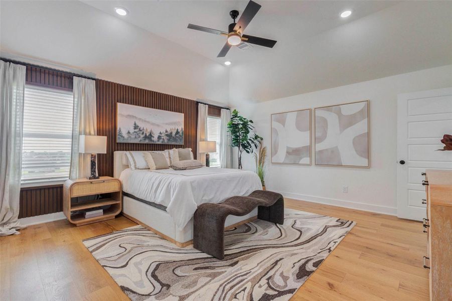 This serene primary bedroom retreat features soaring ceilings, warm wood flooring, and a striking accent wall that adds texture and depth. Large windows invite in natural light, while soft neutral tones create a calming atmosphere. A thoughtfully designed layout offers both comfort and style, with ample space for relaxation and personalization. The picture shown is from the model home with the same floor plan, Rev floor plan.