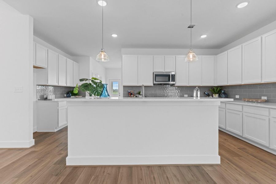 Chef-inspired kitchen boasts 42" white shaker-style cabinetry, gleaming quartz countertops, subway-patterned ceramic tile backsplash, deep undermount sink, and stainless appliances—all centered around a wide island with pendant lighting and bar seating. **Image Representative of Plan Only and May Vary as Built**.
