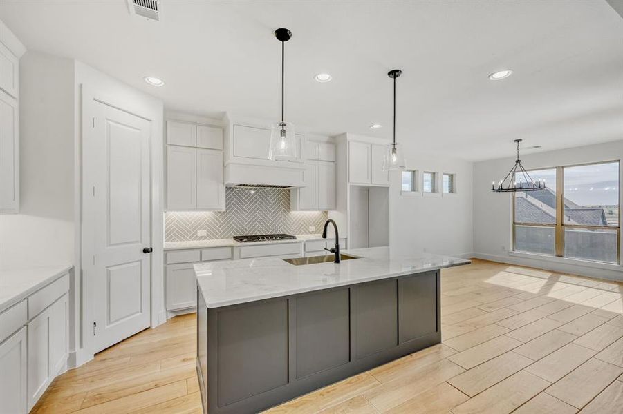 Kitchen featuring light wood-type flooring, dual tone cabinetry, light stone countertops, a center island with sink, and suspended lighting