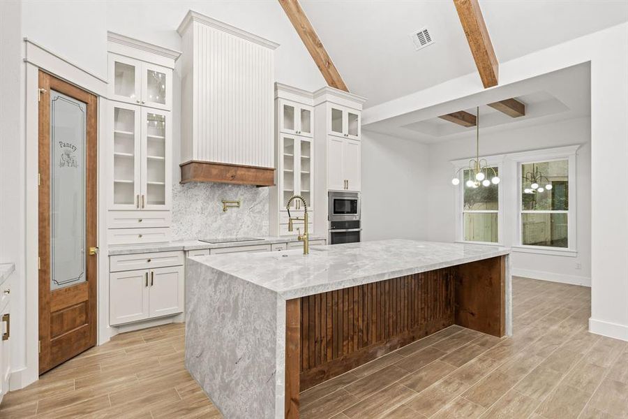 Spacious kitchen featuring a large waterfall island with a wood-paneled base, wood-finish tile flooring, exposed wood ceiling beams, and white cabinetry with glass-front uppers