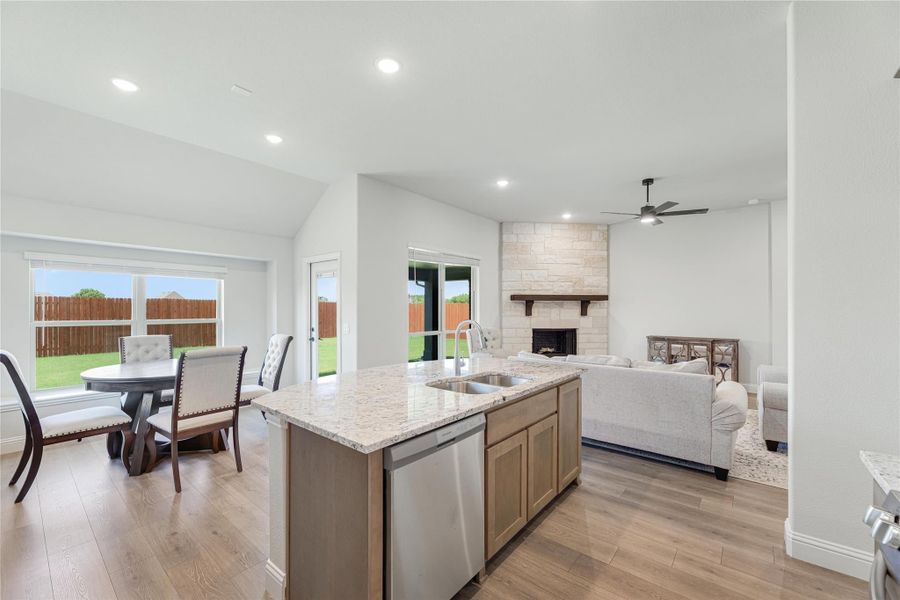Open-concept kitchen with granite island, flowing into dining area and living room with stone fireplace and hardwood floors.