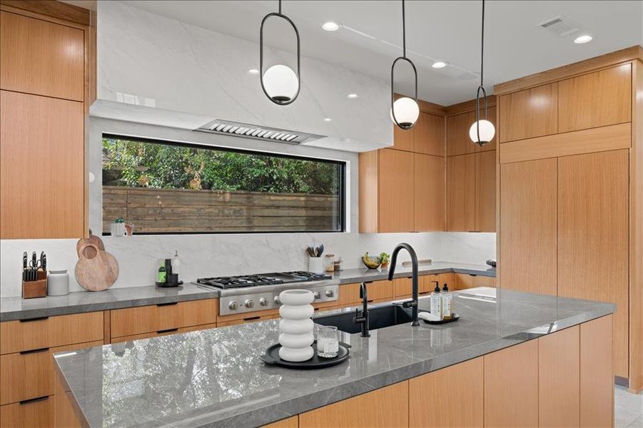 Kitchen featuring modern cabinets, decorative light fixtures, dark stone counters, stainless steel gas cooktop, and a kitchen island with sink