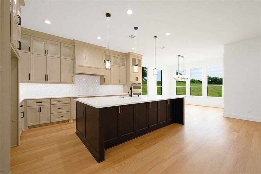 Kitchen featuring a kitchen island with sink, two tone cabinetry, glass fronted cabinets, light wood-style flooring, and pendant lighting