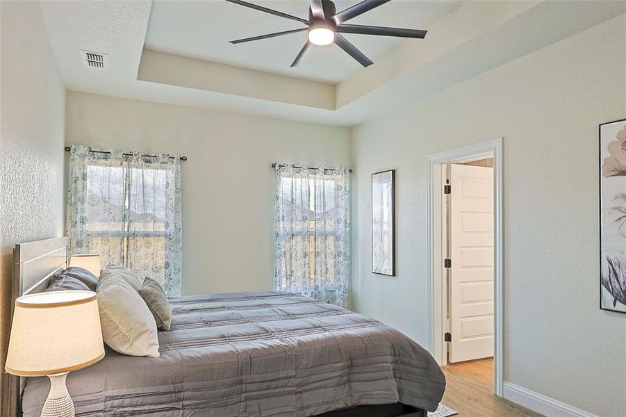Room featuring a tray ceiling with a ceiling fan, two windows with sheer curtains, a five-panel interior door, and wood-finish flooring