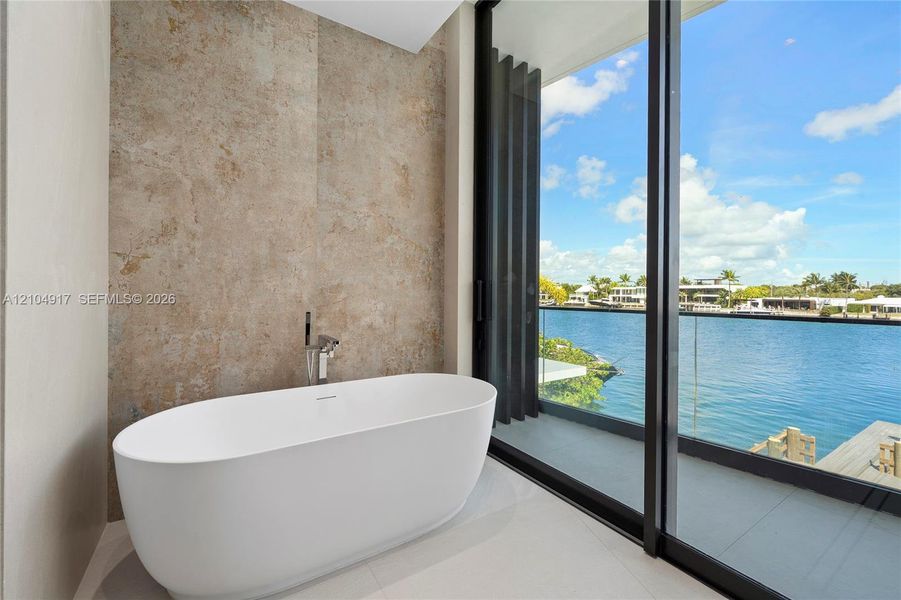 Freestanding bathtub with a floor-mounted faucet, textured wall tile, and full-height sliding glass doors opening to a waterfront balcony