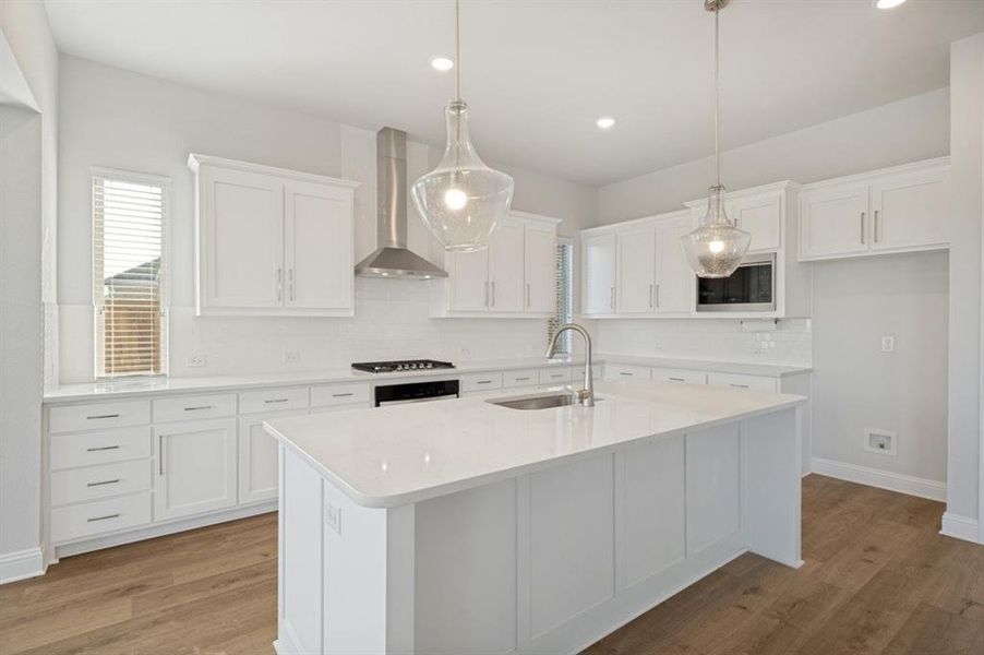 Bright kitchen featuring a central island with an undermount sink, white cabinetry, integrated stainless steel appliances, pendant lighting, and wood-finish flooring