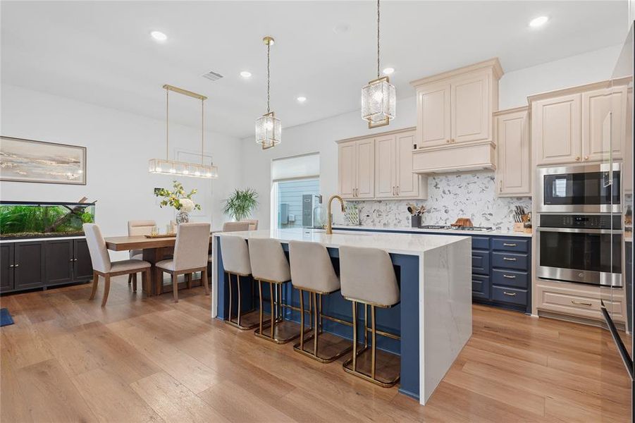 Open-concept kitchen featuring a waterfall island with seating, stainless steel appliances, cream and navy cabinetry, light wood-finish flooring, and a patterned tile backsplash Open-concept kitchen featuring a waterfall island with seating, stainless steel appliances, cream and navy cabinetry, light wood-finish flooring, and a patterned tile backsplash