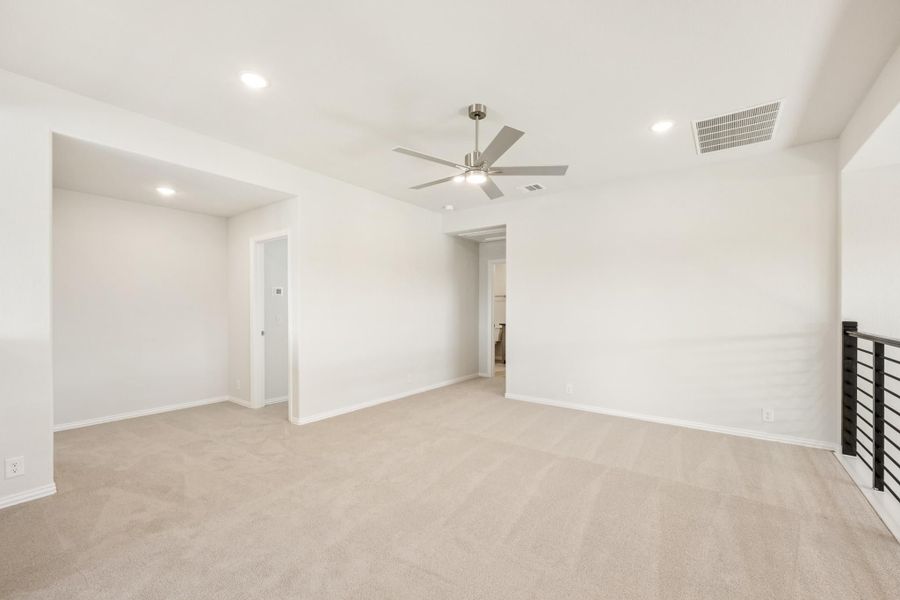 Spacious empty room with beige carpet, ceiling fan, white walls, and metal stair railing Spacious empty room with beige carpet, ceiling fan, white walls, and metal stair railing