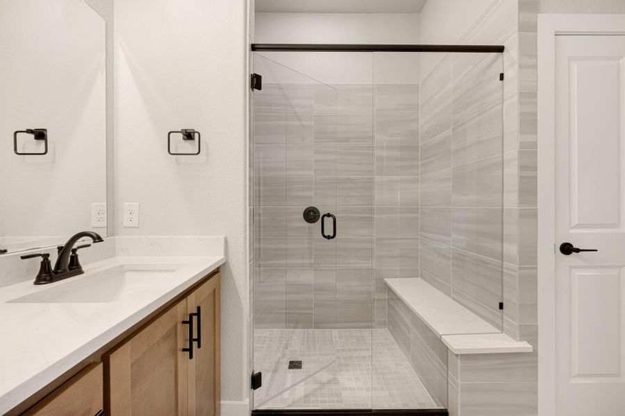 Bathroom with glass-door walk-in shower, gray tile walls, built-in bench, and wood vanity with white countertop Bathroom with glass-door walk-in shower, gray tile walls, built-in bench, and wood vanity with white countertop