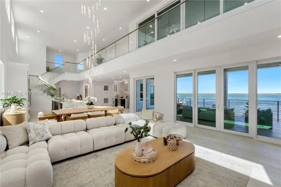 Expansive open-concept living area featuring double-height ceilings, a floor-to-ceiling glass-paneled mezzanine, multi-panel sliding glass doors, and a linear pendant light fixture Expansive open-concept living area featuring double-height ceilings, a floor-to-ceiling glass-paneled mezzanine, multi-panel sliding glass doors, and a linear pendant light fixture