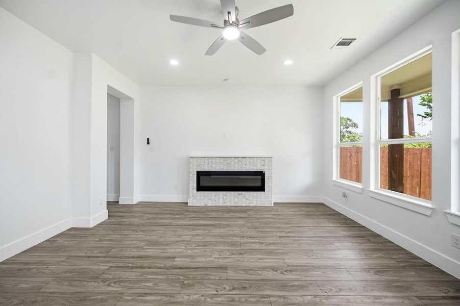 Spacious room featuring wood-finish flooring, a modern linear fireplace with a mosaic tile surround, multiple large windows, a ceiling fan with integrated lighting, and recessed ceiling lights Spacious room featuring wood-finish flooring, a modern linear fireplace with a mosaic tile surround, multiple large windows, a ceiling fan with integrated lighting, and recessed ceiling lights